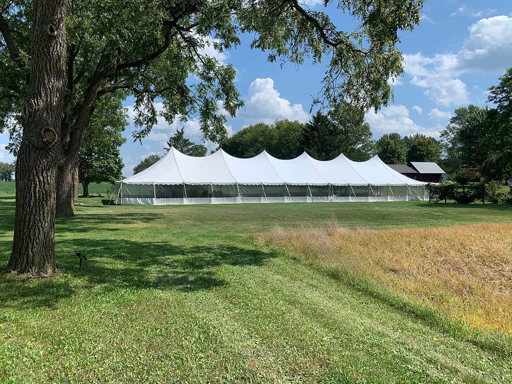 A large white event tent stands on a sunny, grassy field surrounded by trees. The sky above is partly cloudy.