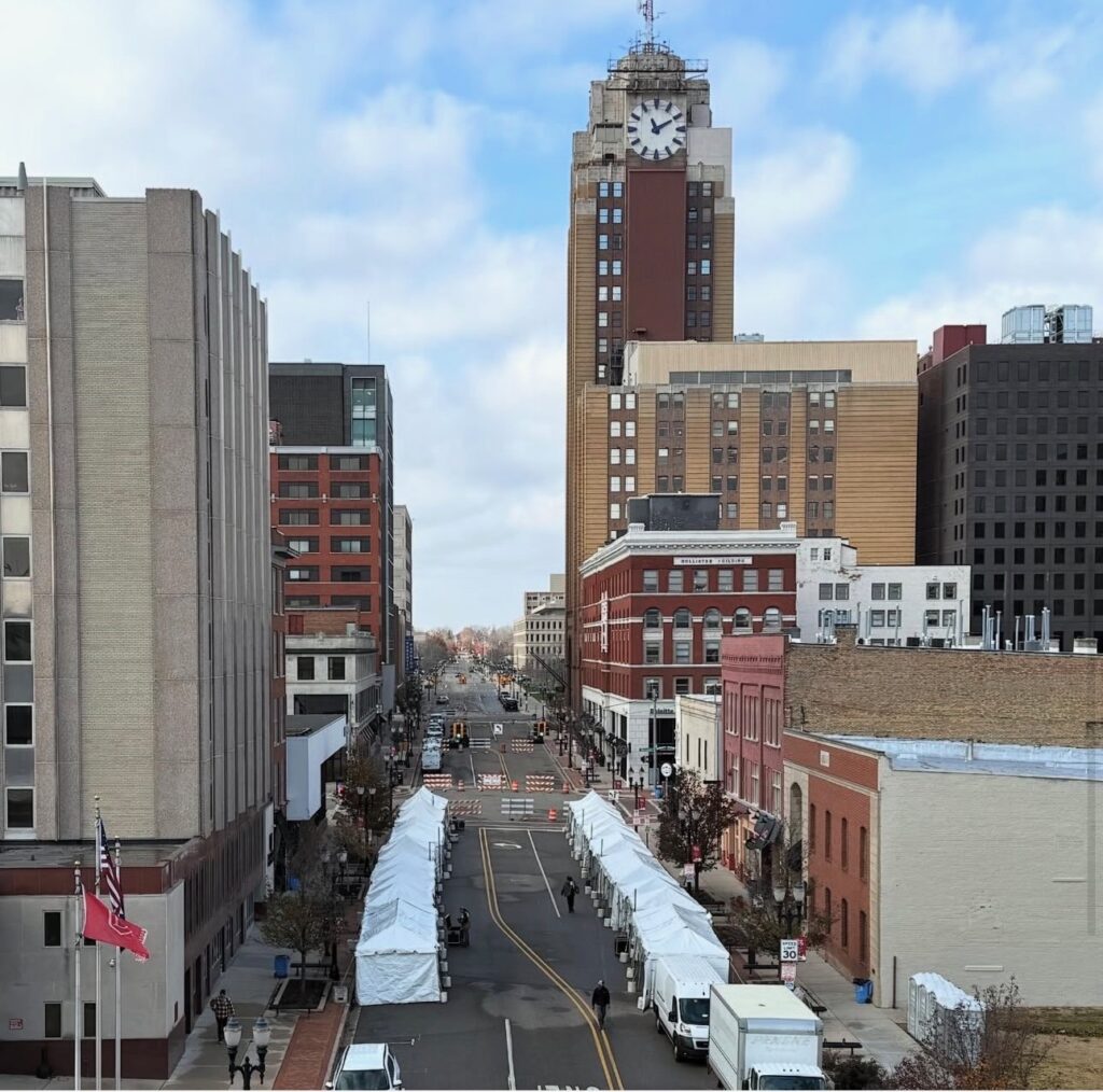 An urban scene with a street lined with empty white tents for a holiday festival. Tall buildings surround the street, with a prominent clock tower under a cloudy sky.