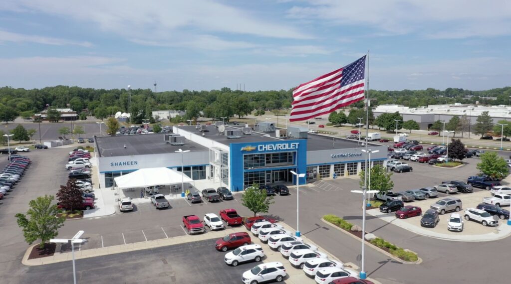 Aerial view of a Chevrolet dealership with a large American flag and a tent assembled out front. The parking lot is filled with parked cars. The building is surrounded by trees and neighboring structures under a partly cloudy sky.