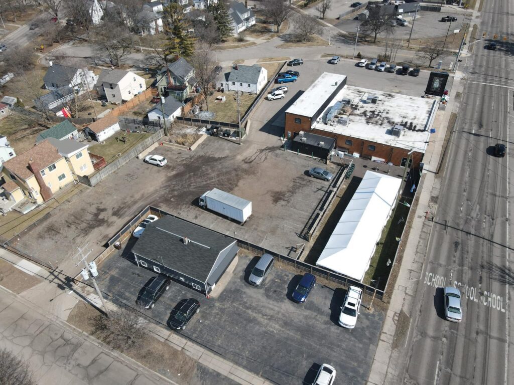 Aerial view of a commercial lot with a black-roofed building, several parked cars, and a large white tent. Surrounding the lot are small residential houses.