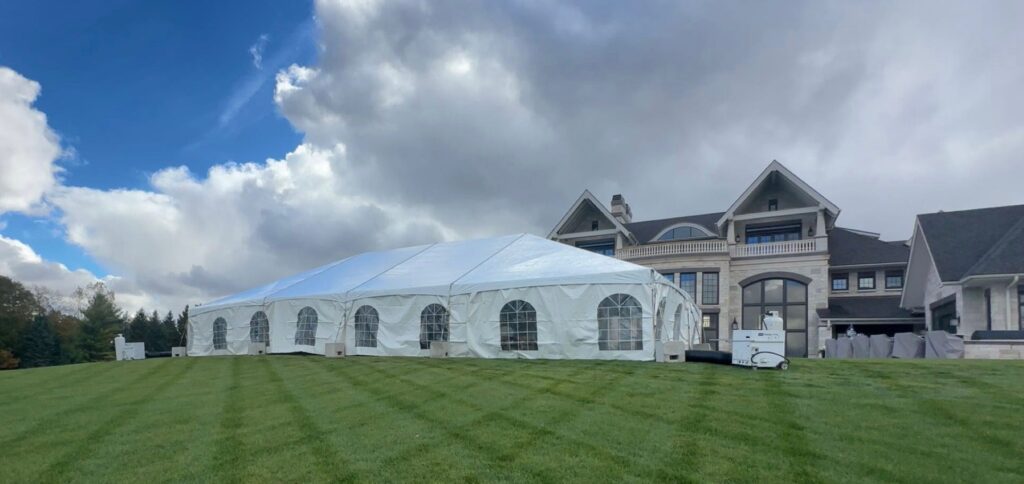 Large white event tent on a lush green lawn, set against a large house with a cloudy blue sky backdrop.