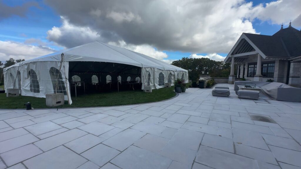 A large white event tent stands on a grassy area next to a spacious stone patio with a partly cloudy sky overhead. Nearby is a house with a classic roof.