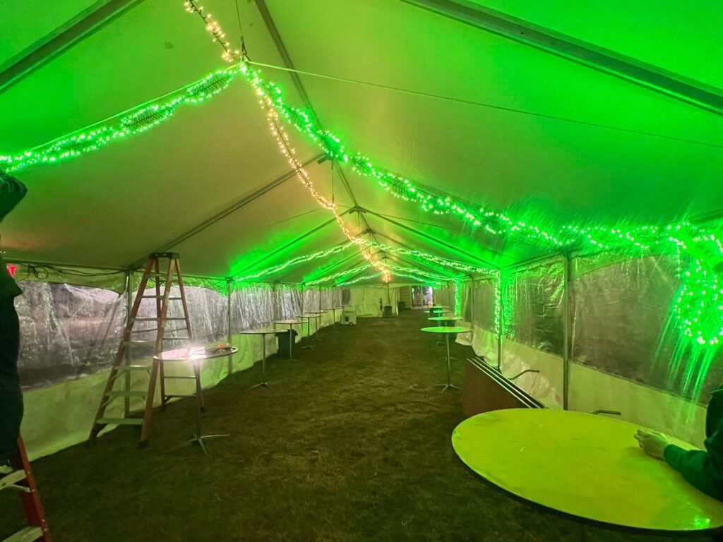A long event tent with green LED lights strung along the ceiling, creating a festive atmosphere. Several round tables and a wooden ladder are visible inside.