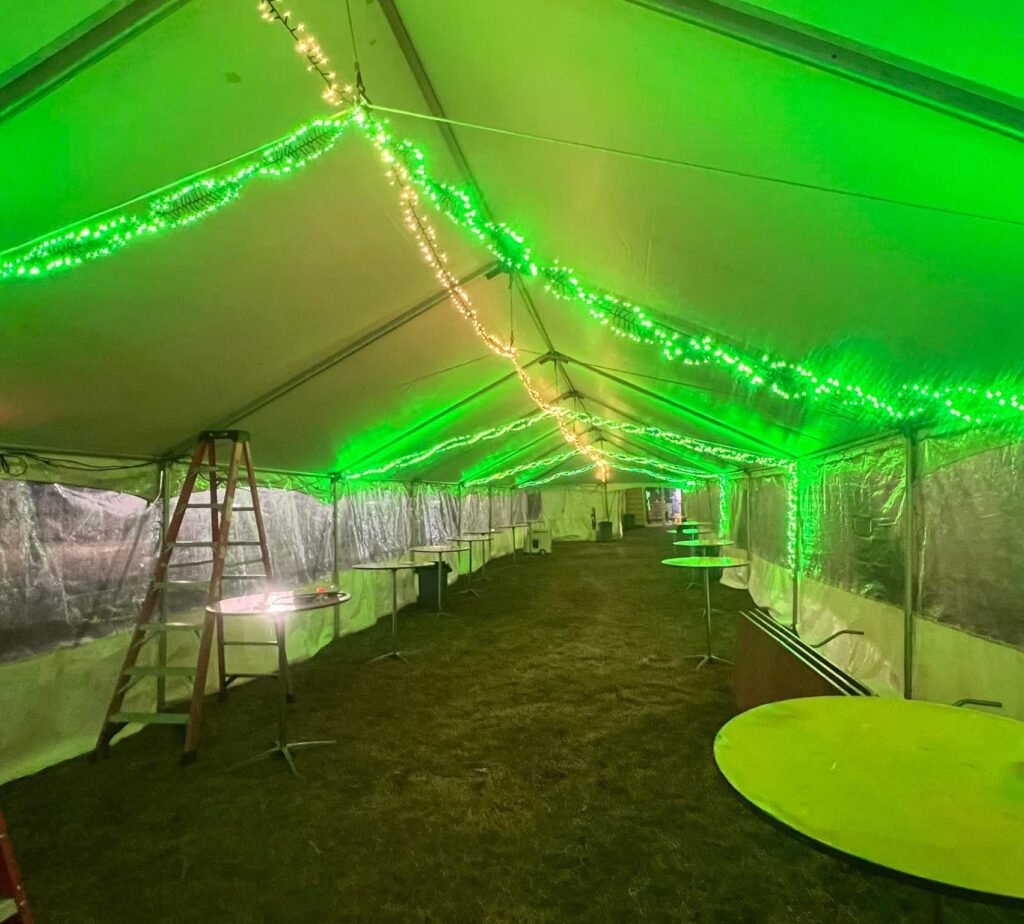 A long event tent with green LED lights strung along the ceiling, creating a festive atmosphere. Several round tables and a wooden ladder are visible inside.