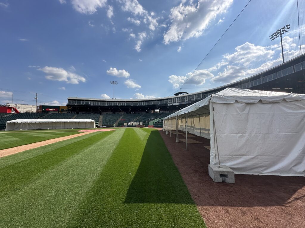 Wide view of an empty lansing lugnuts baseball stadium under a bright blue sky. Tents are set up on the right side of the green outfield.