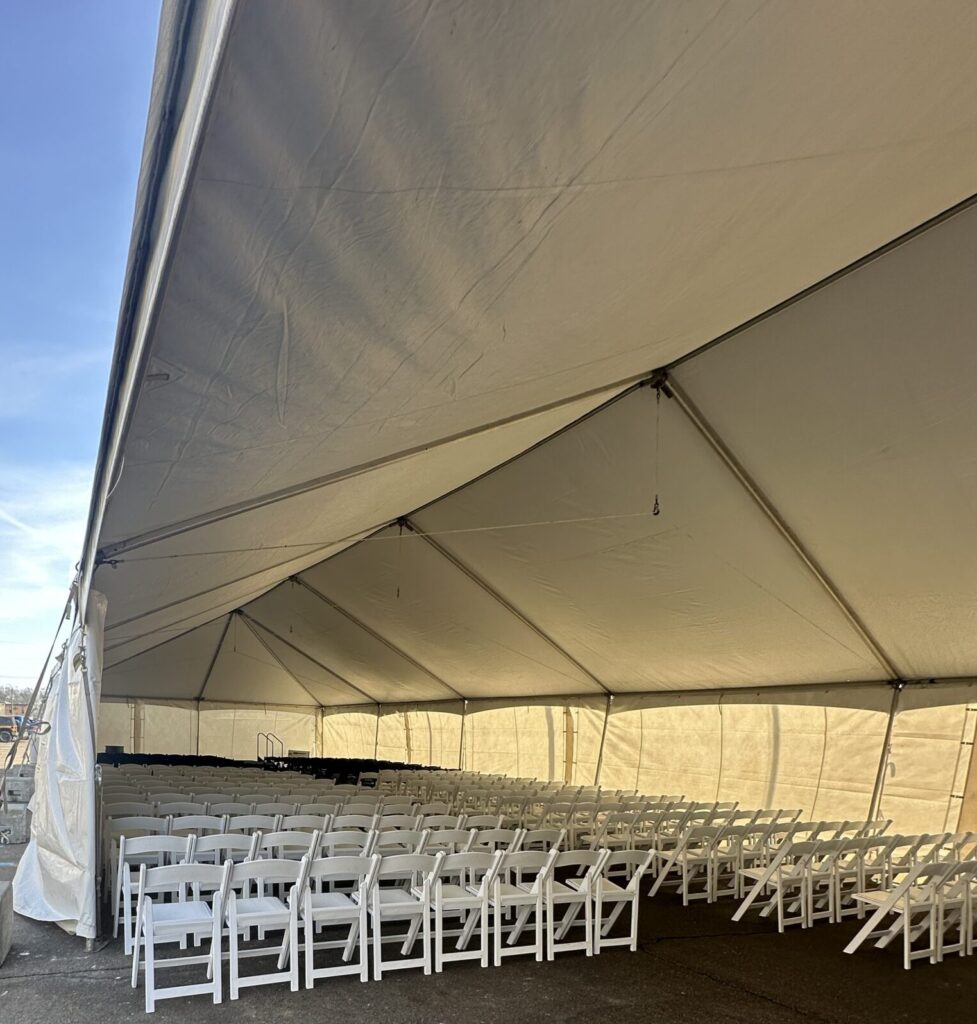 An empty outdoor tent with rows of white folding chairs on a paved surface, under a clear blue sky.