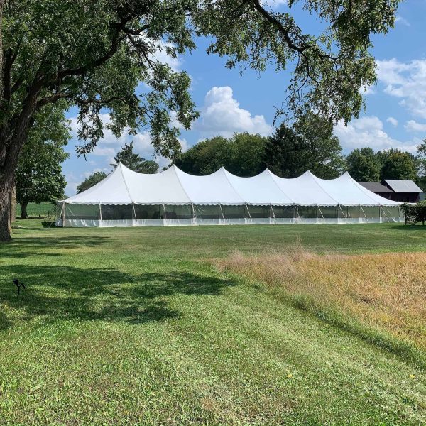 A large white event tent stands on a sunny, grassy field surrounded by trees. The sky above is partly cloudy.