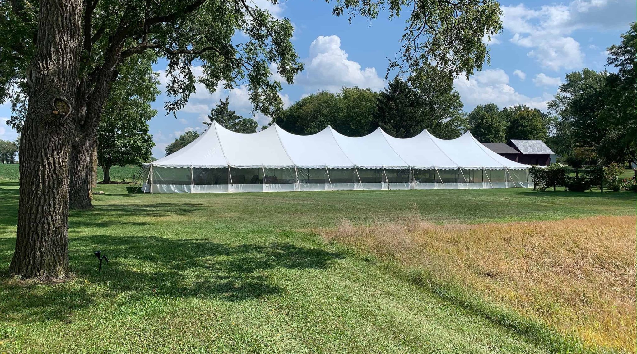 A large white event tent stands on a sunny, grassy field surrounded by trees. The sky above is partly cloudy.